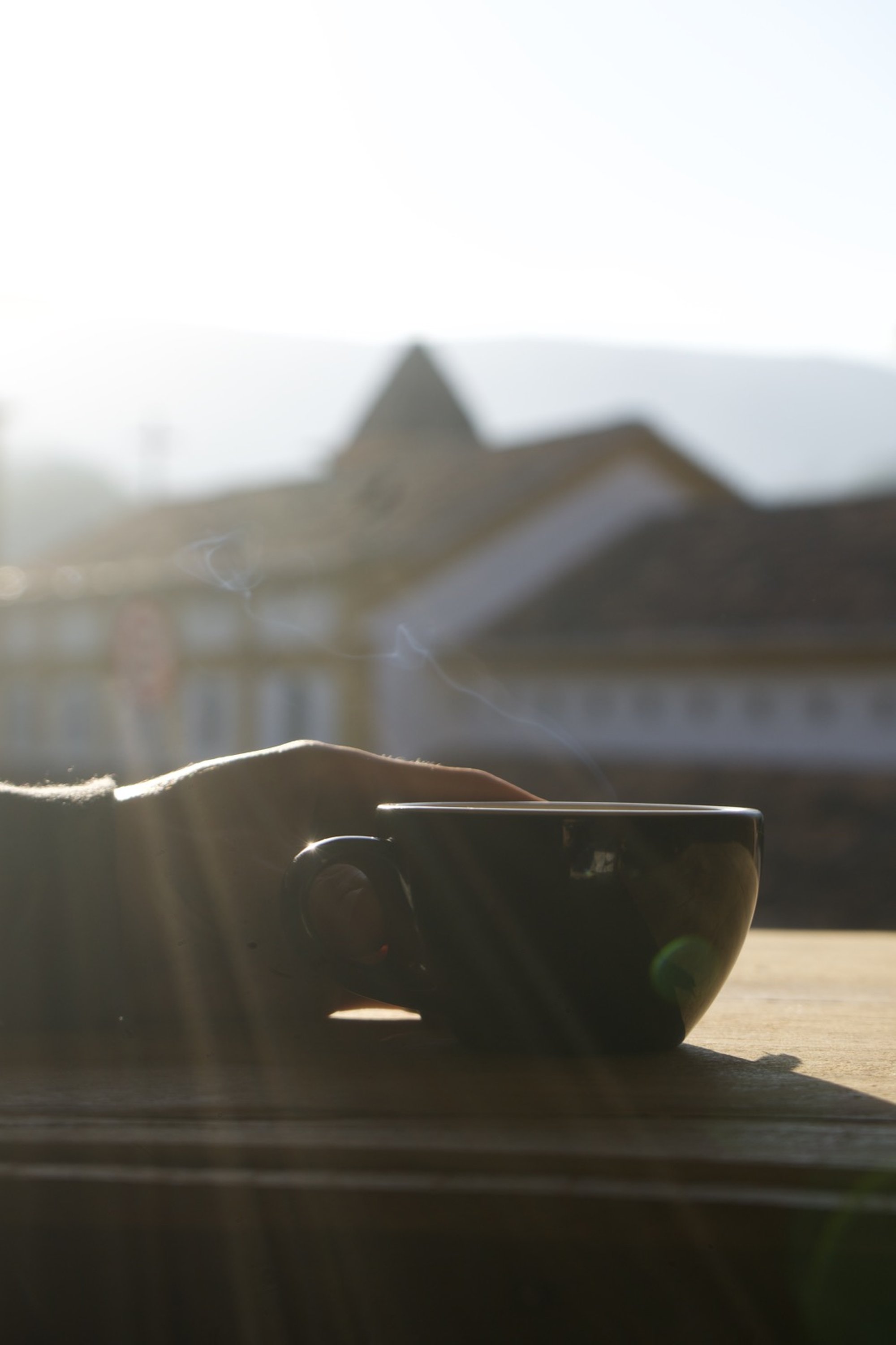 Mão segurando uma xícara de café fumegante ao amanhecer, com a igreja de Piedade do Paraopeba ao fundo